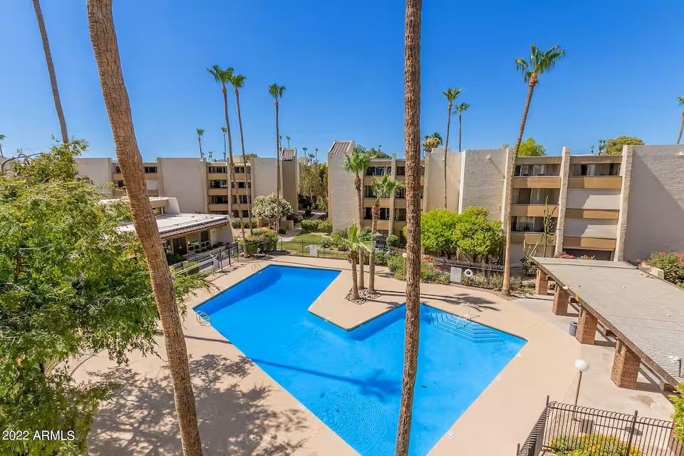 An L-shaped swimming pool in a courtyard surrounded by multi-story tan apartment buildings and tall palm trees.