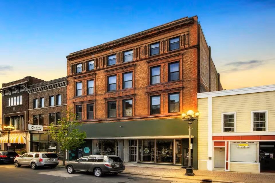 A street view of a four-story red brick commercial building between a smaller brick building and a light-colored storefront.