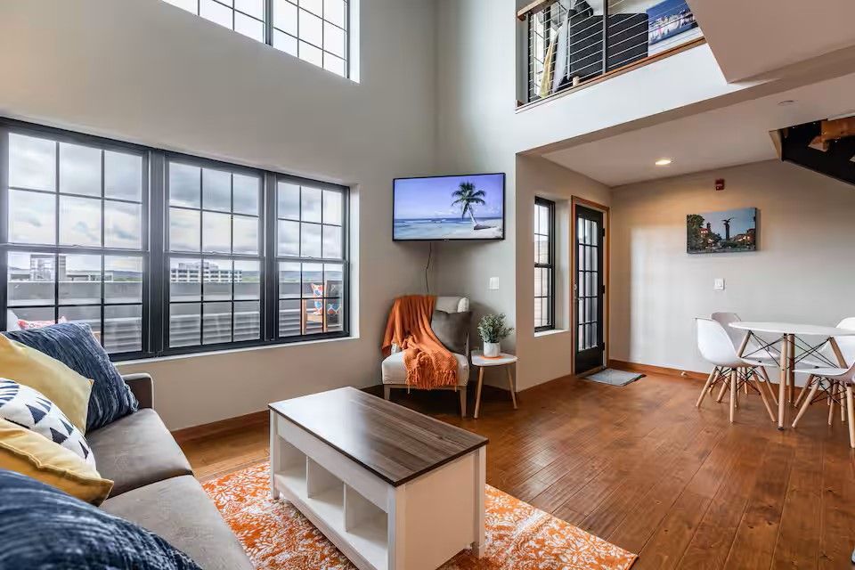 A bright living area featuring a couch, coffee table, accent chair, and dining set on wood floors with high windows.