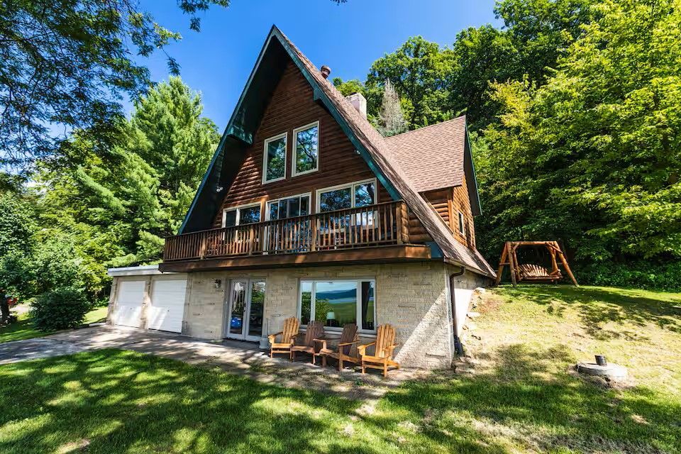 A rustic A-frame cabin with a wooden upper level and stone base, surrounded by trees with a swing set on a grassy hill.