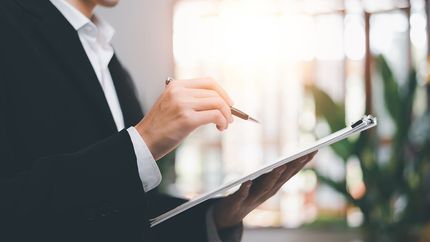 A man in a suit is holding a clipboard and pointing at it with a pen.