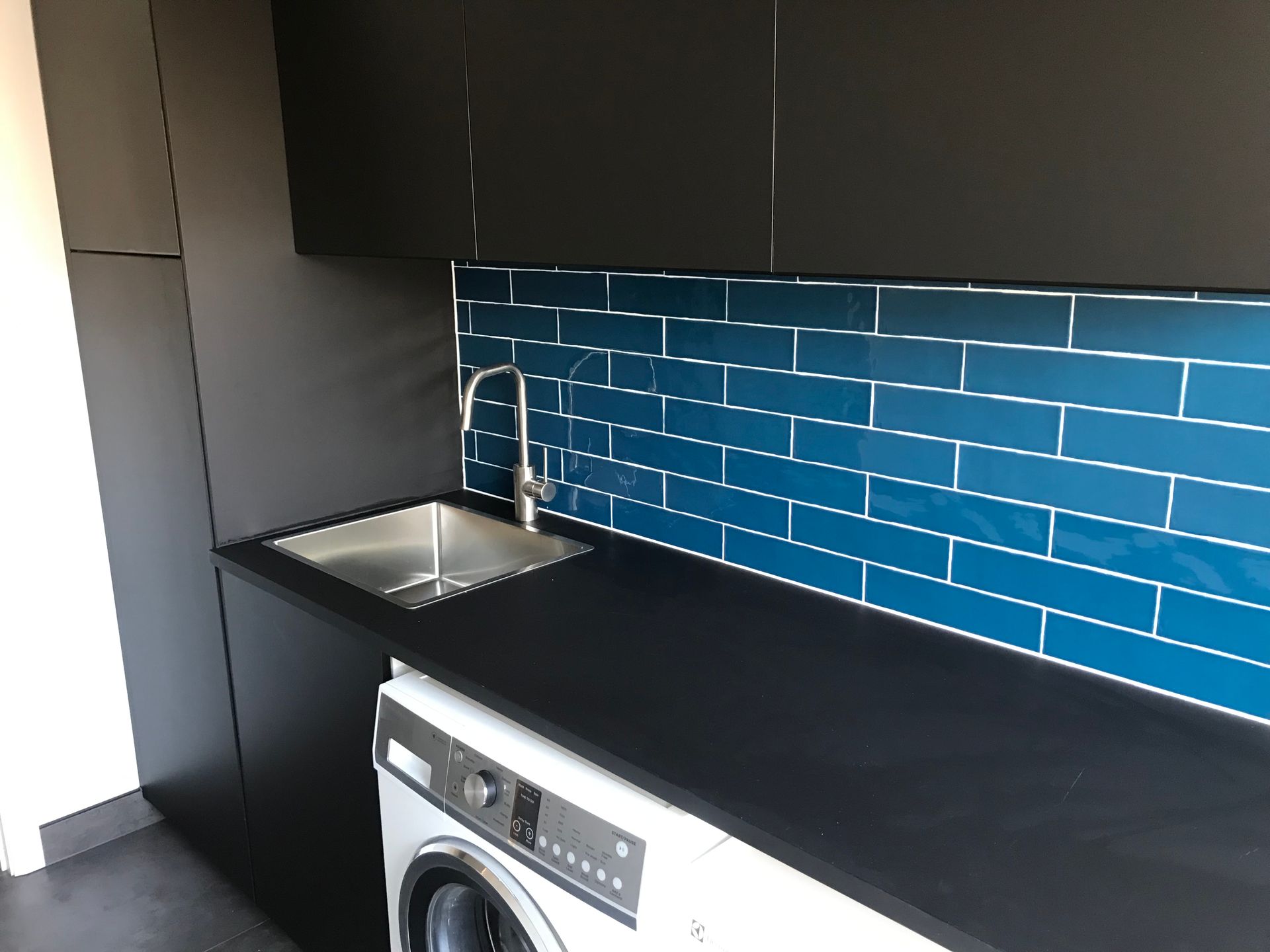 Laundry room with a washing machine, sink, and blue tiled backsplash. Black cabinetry and countertop. — Rectified Edge Tiling & Bathroom Renovations in Queanbeyan, NSW