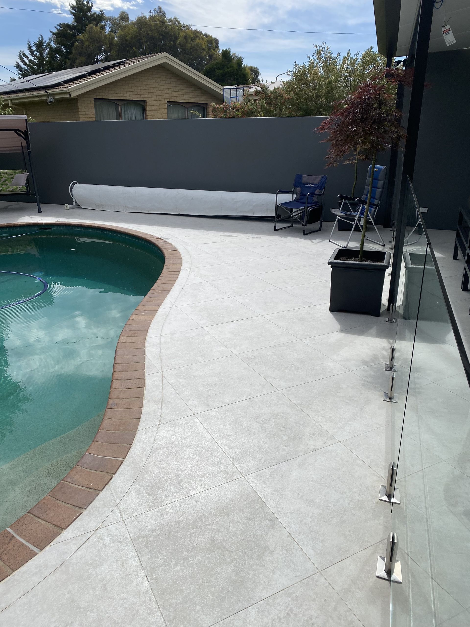 Poolside concrete patio with a pool on the left and a dark fence in the background. A small tree sits in a pot. — Rectified Edge Tiling & Bathroom Renovations in Queanbeyan, NSW