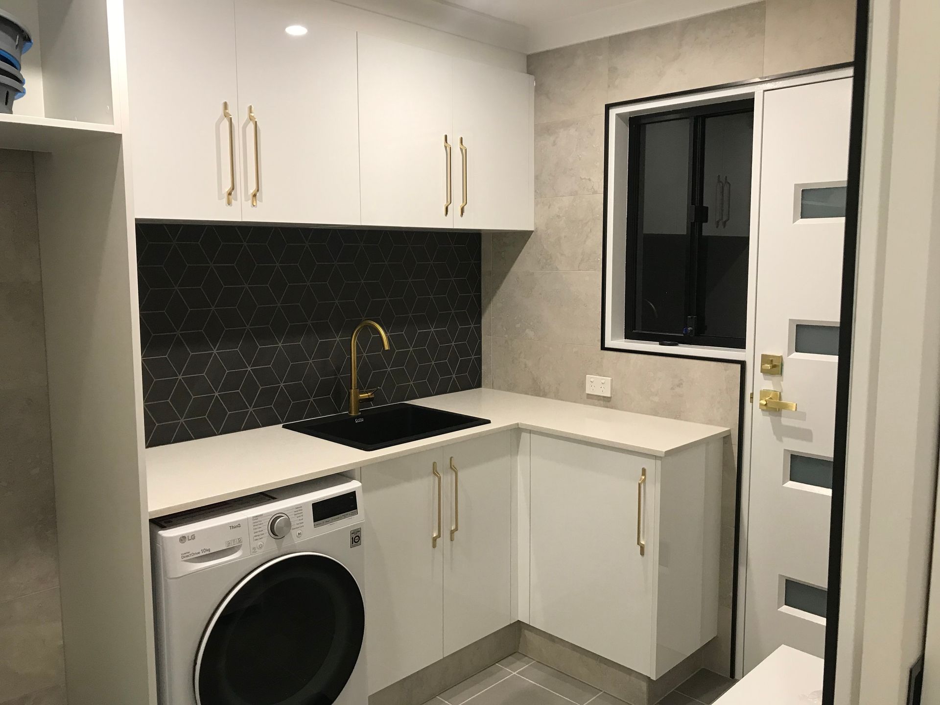 Laundry room with white cabinets, black sink and backsplash, washer, gold faucet and hardware, and a window. — Rectified Edge Tiling & Bathroom Renovations in Fyshwick, ACT