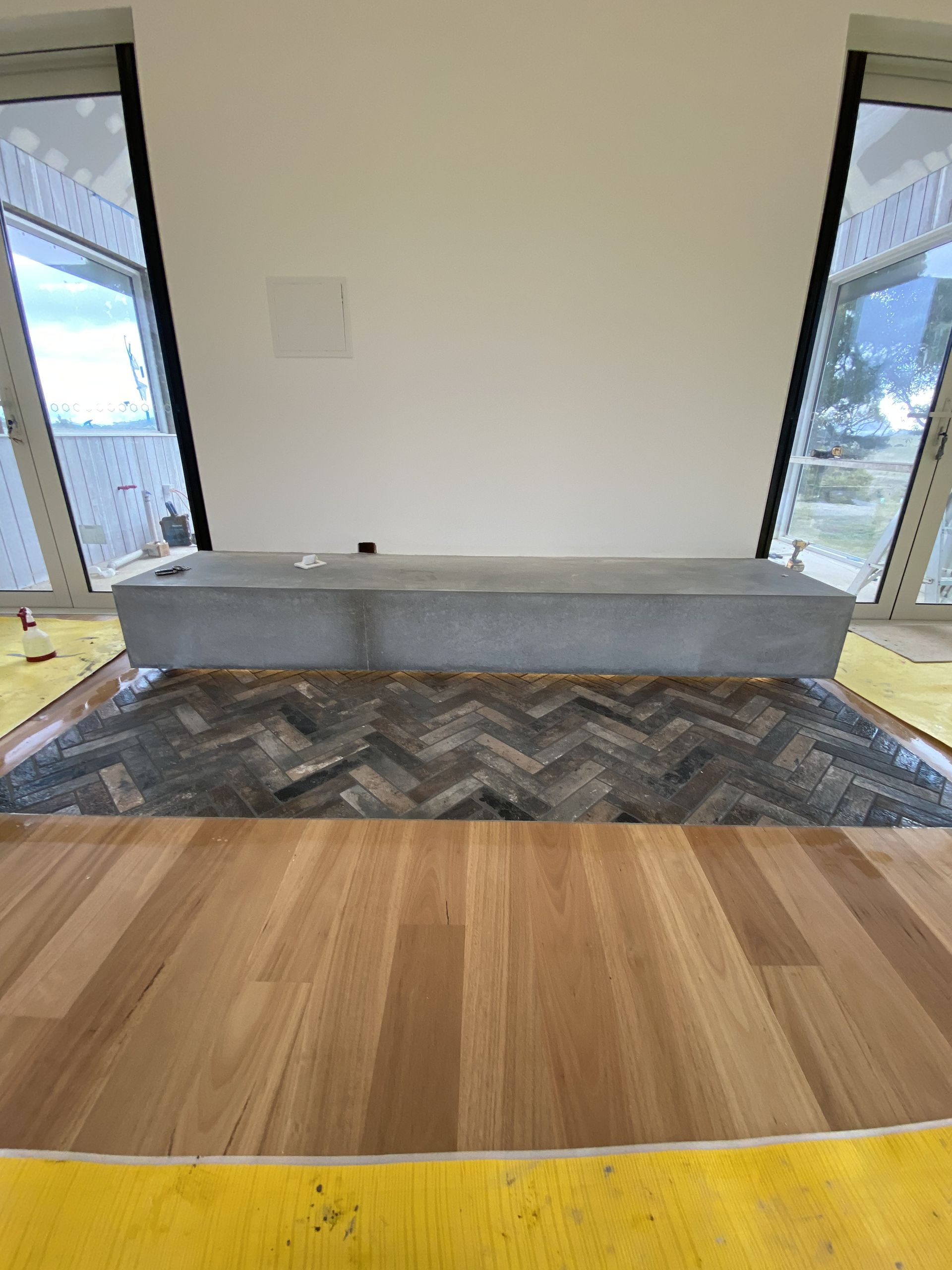 Room with herringbone tile floor, wooden floor, and grey rectangular structure against white wall. — Rectified Edge Tiling & Bathroom Renovations in Batemans Bay, NSW