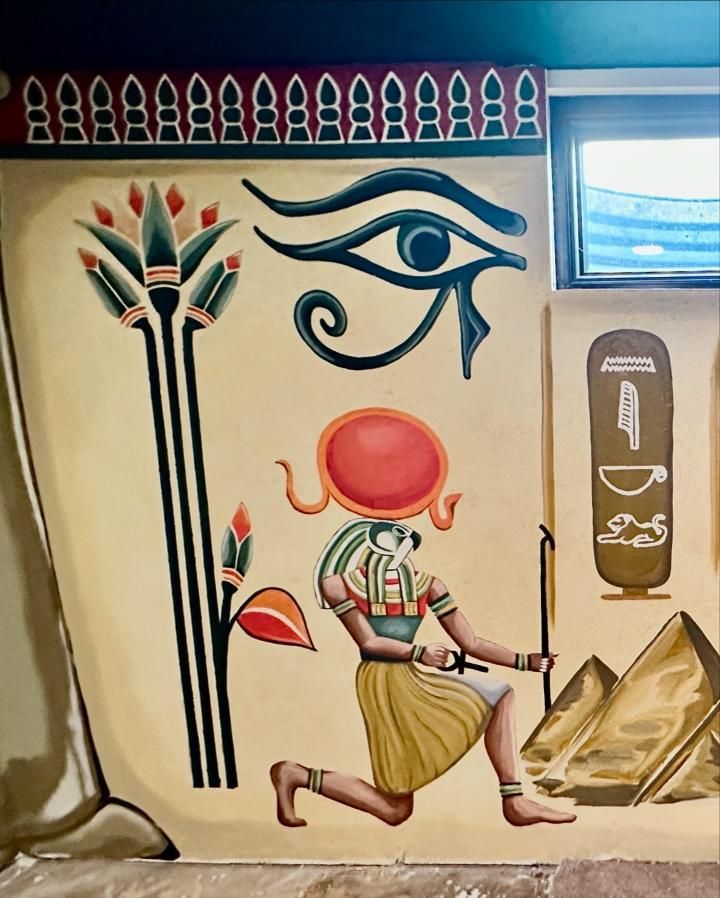 Egyptian-themed mural: lotus, Eye of Horus, deity with sun disc, pyramids, and hieroglyphs on a tan background.