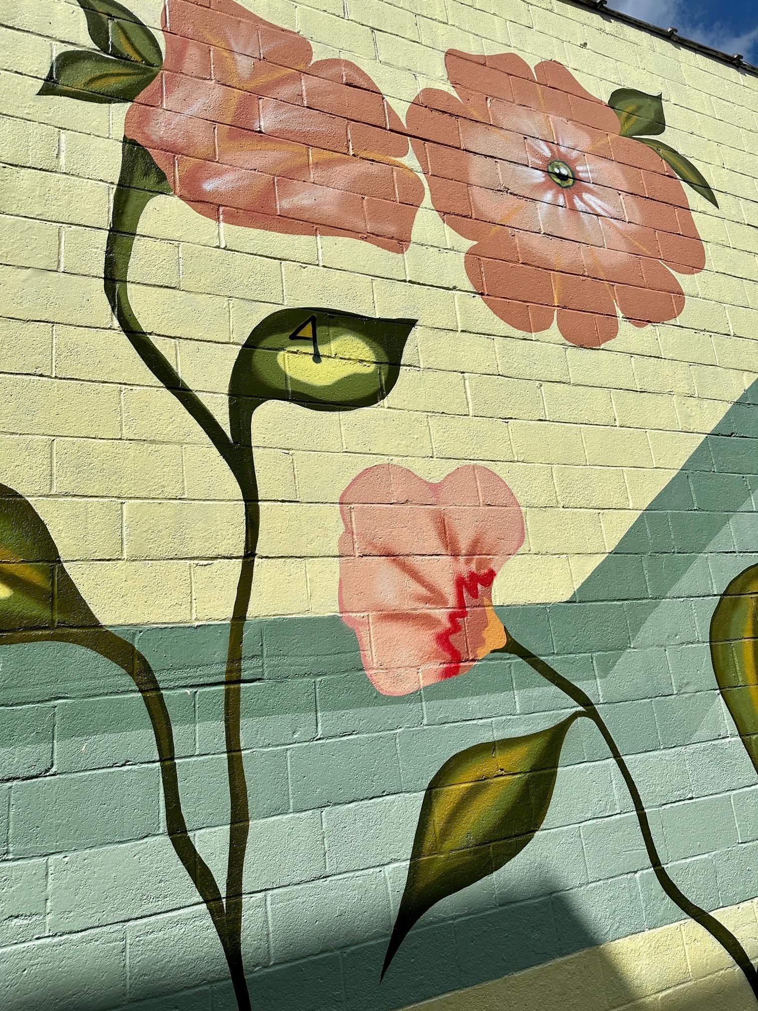 Mural of pink flowers on a brick wall, with green leaves and blue and yellow painted background.