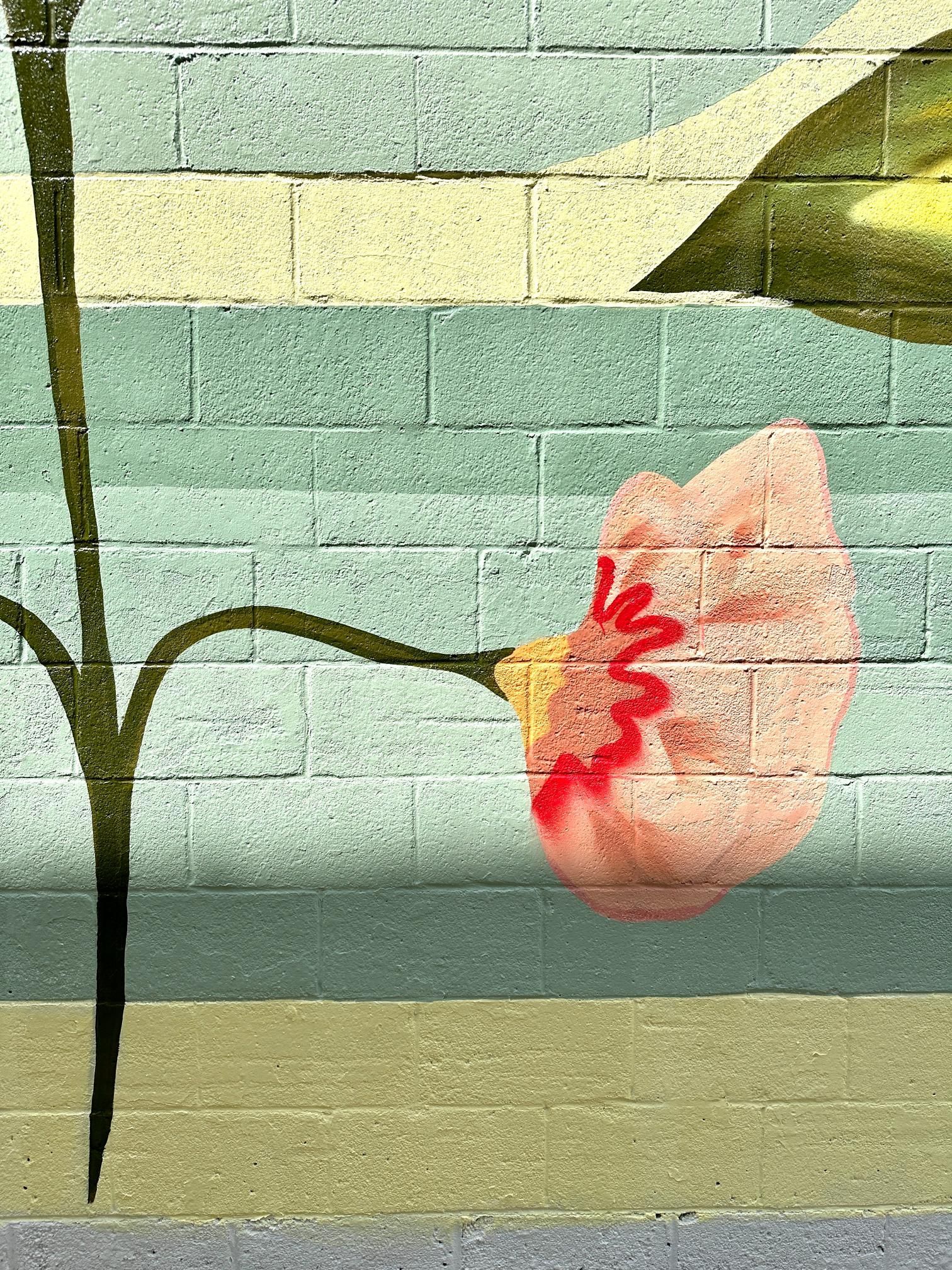Mural of a pink flower with red details, green stem and leaf, on a brick wall with pastel colors.