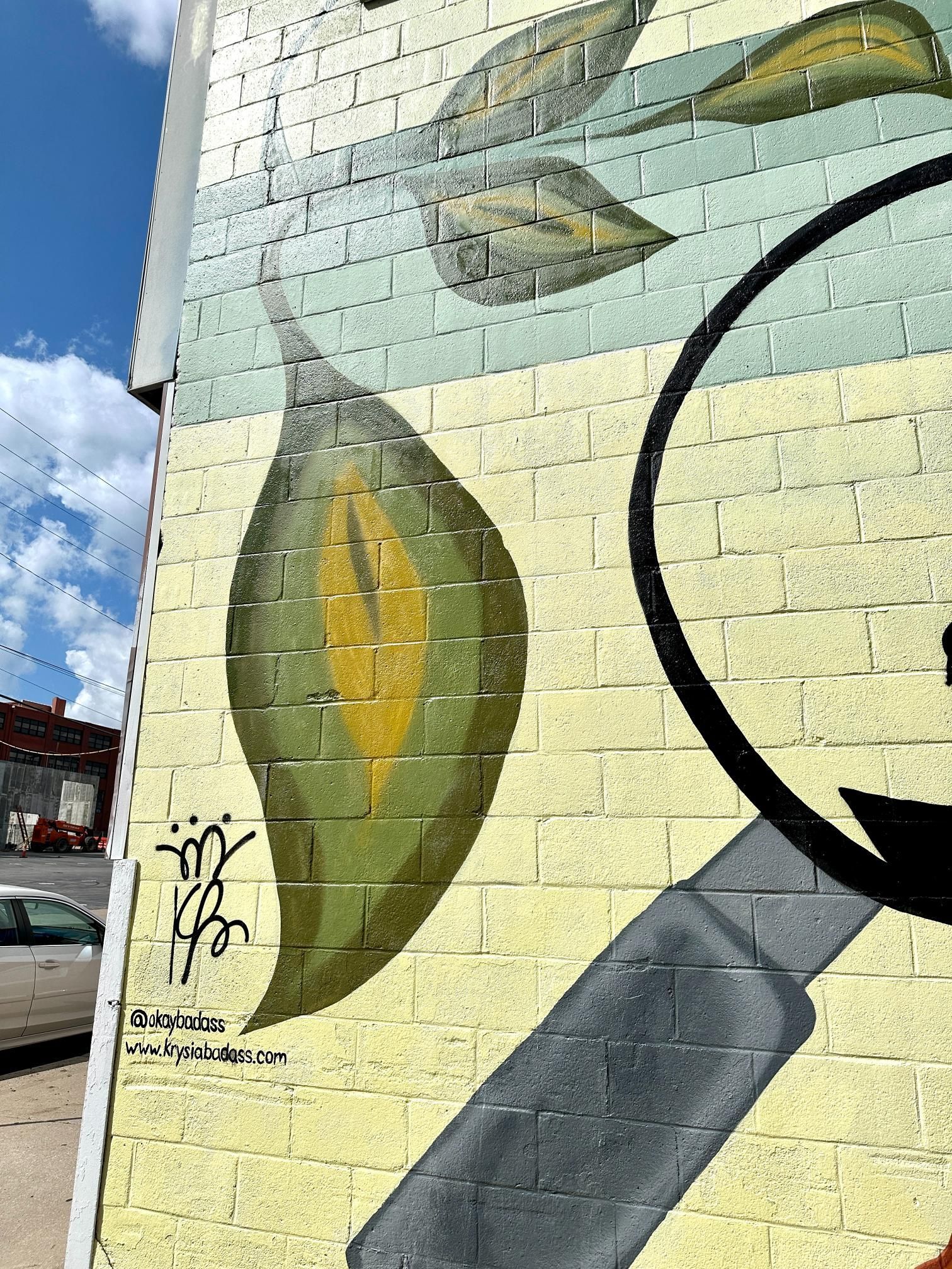 Mural of green leaf and magnifying glass on a brick wall, against a blue sky.