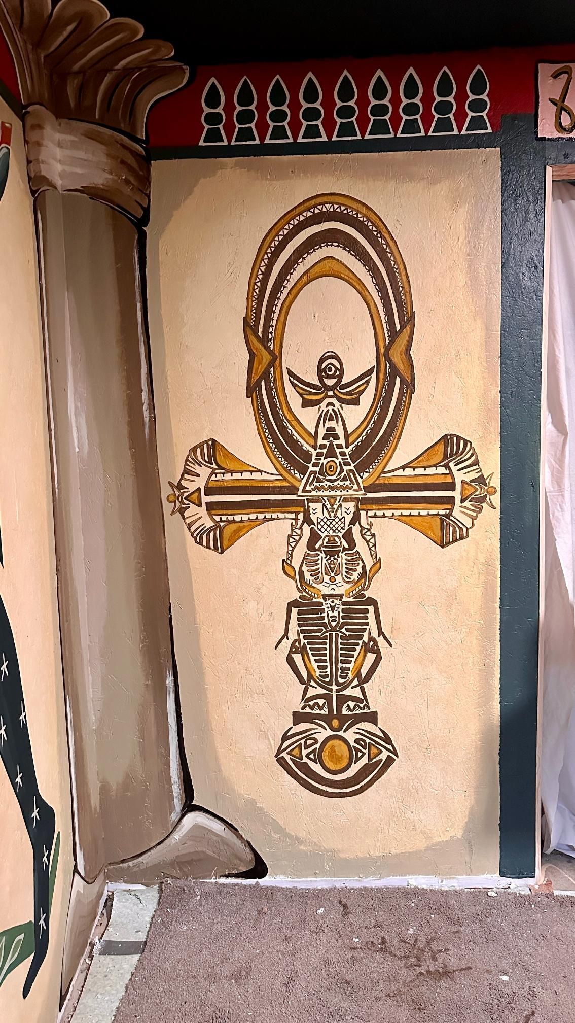 Ankh symbol with intricate detailing, in gold and brown hues, painted on a tan wall.
