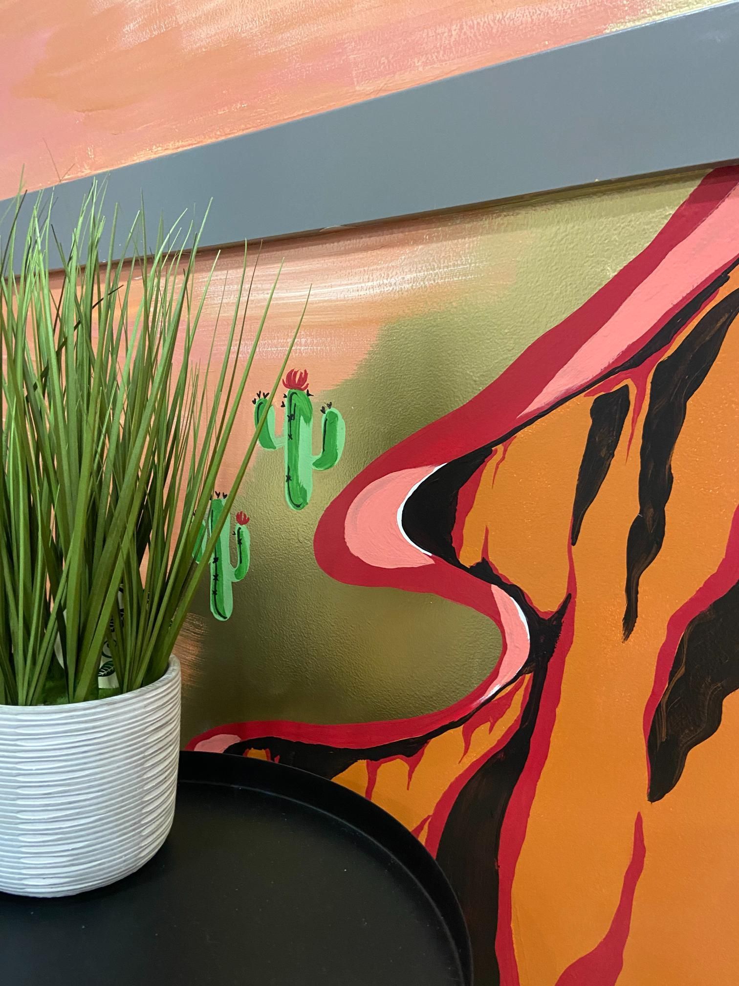 Artificial plant in white pot on black table next to colorful southwestern-style mural.