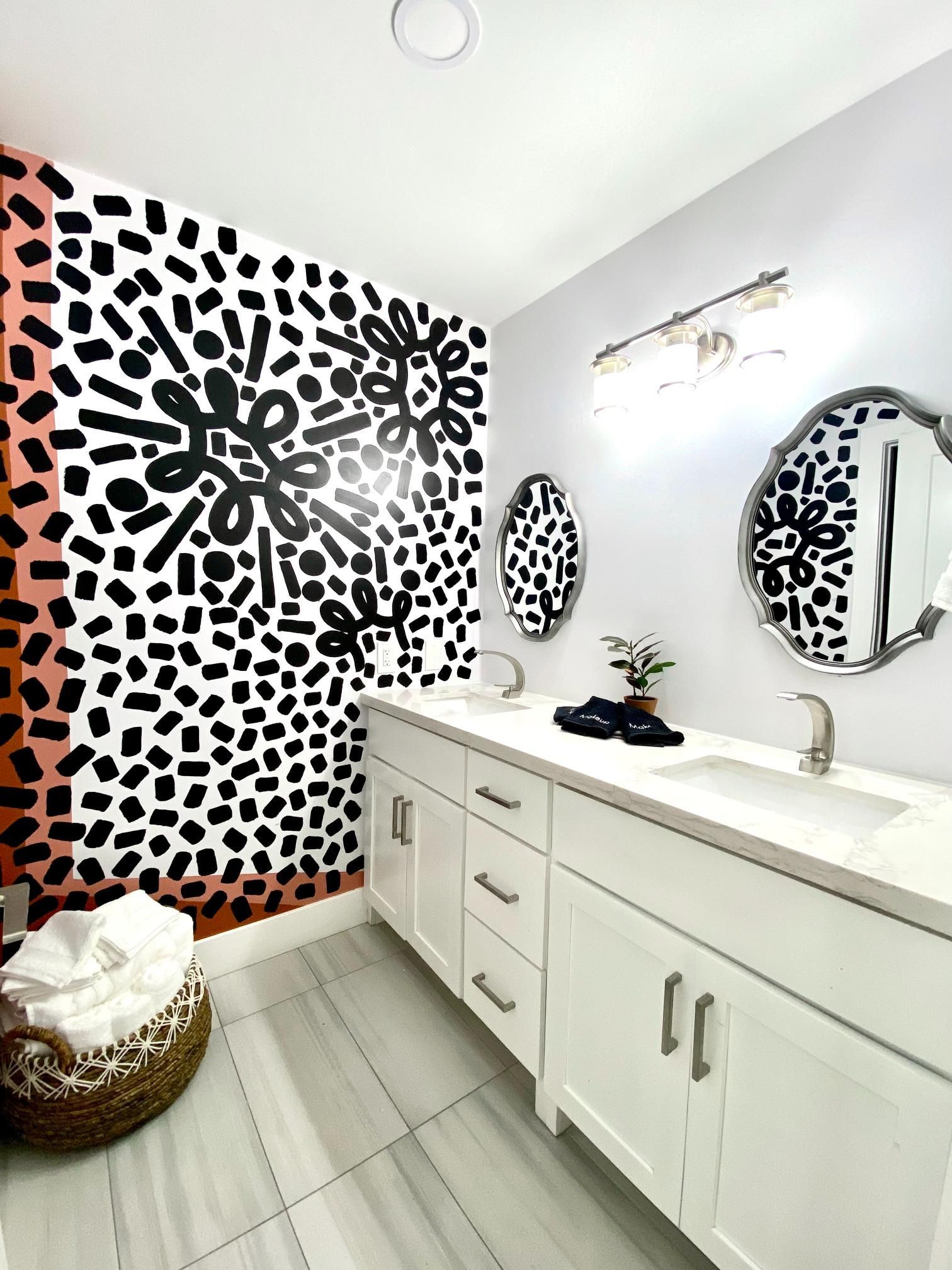 Modern bathroom with white vanity, patterned accent wall, two oval mirrors, and woven basket.