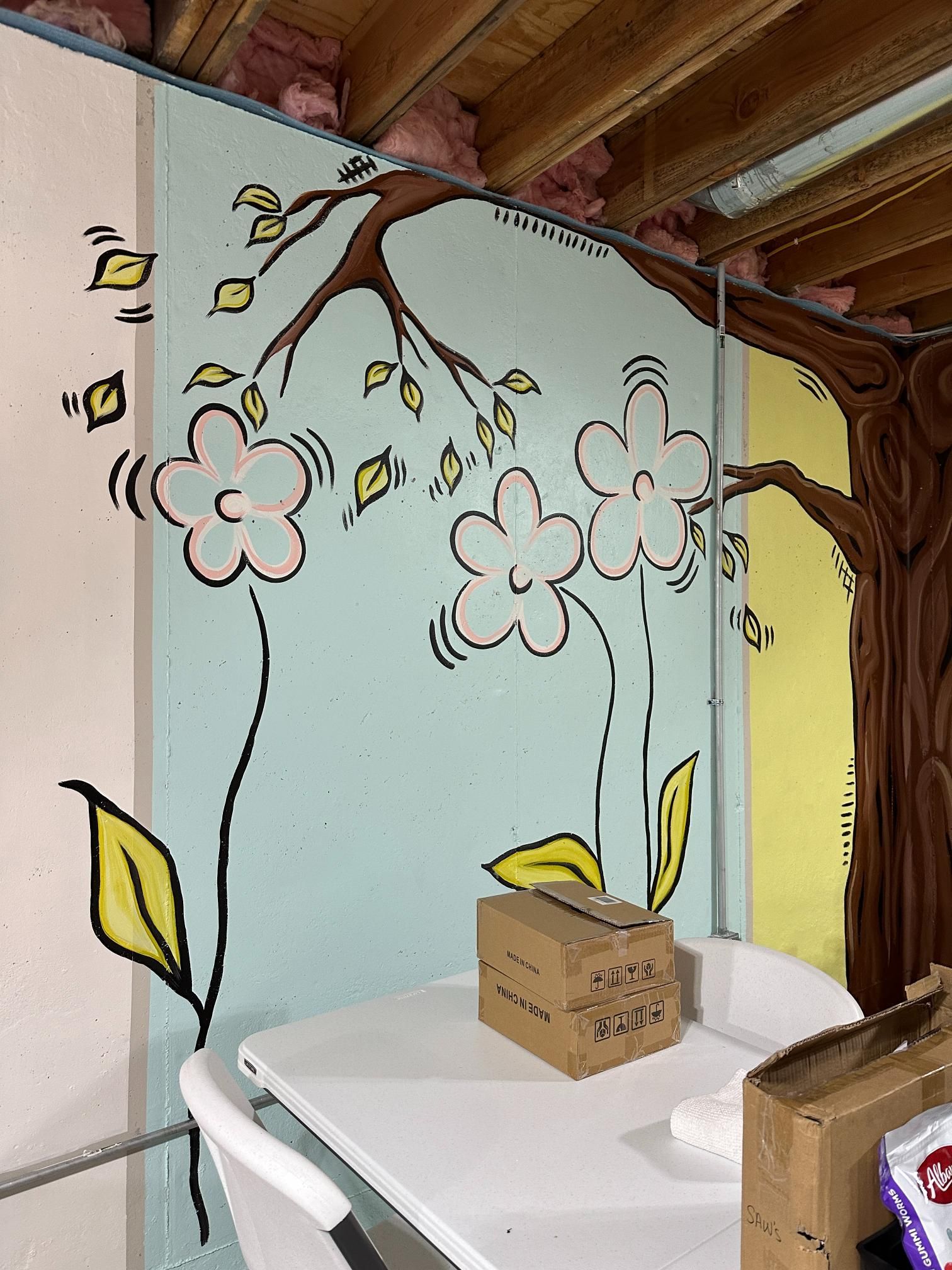 Basement wall mural of flowers and a tree, above a white table with boxes.