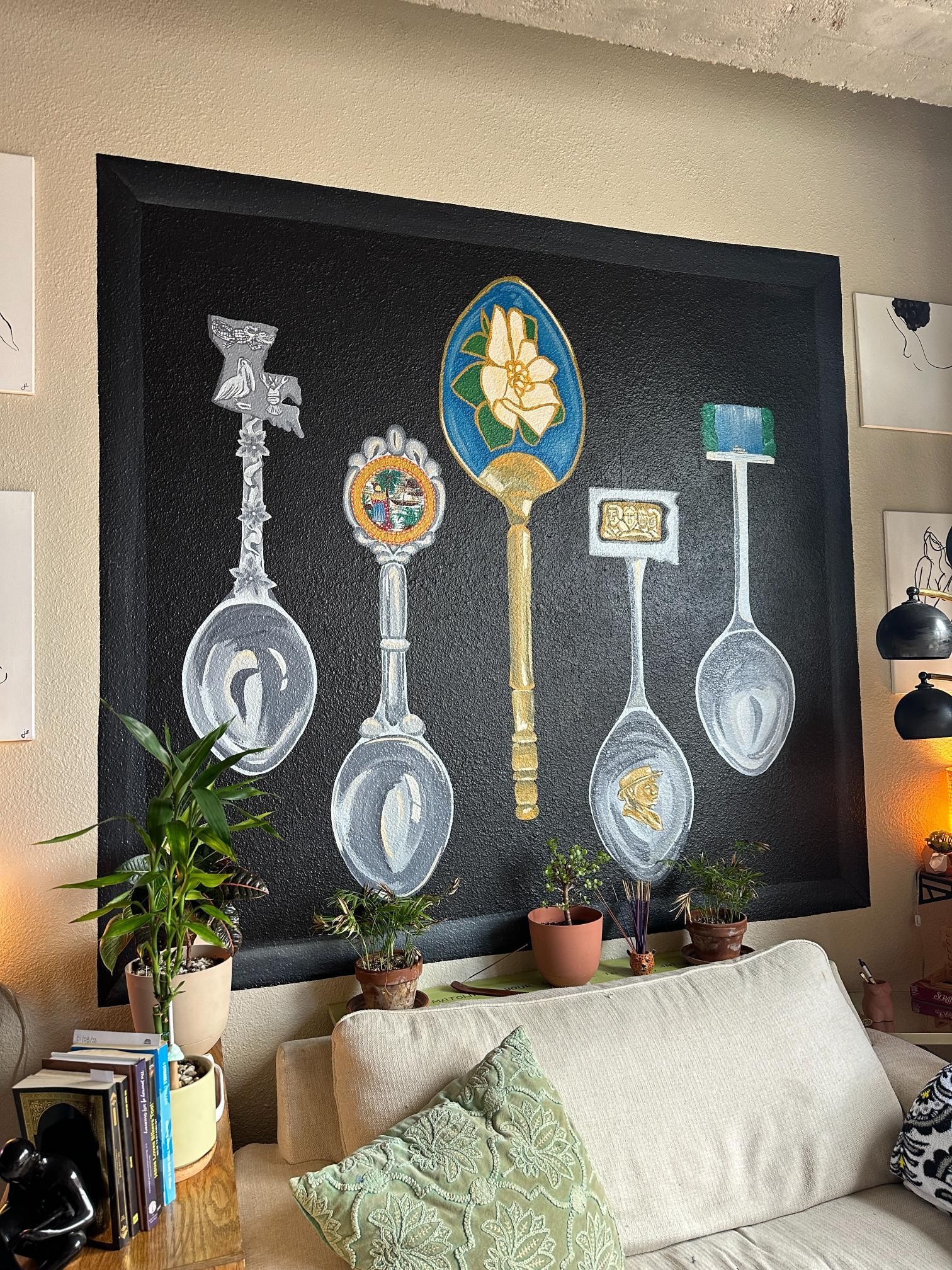Painting of ornate spoons on a black background, mounted above a couch with plants in front.