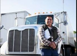 A man is standing in front of a semi truck holding a clipboard.