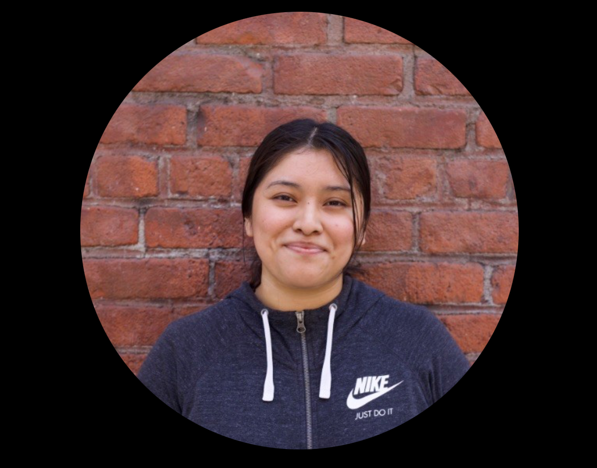 Woman in gray Nike hoodie smiles in front of a brick wall.