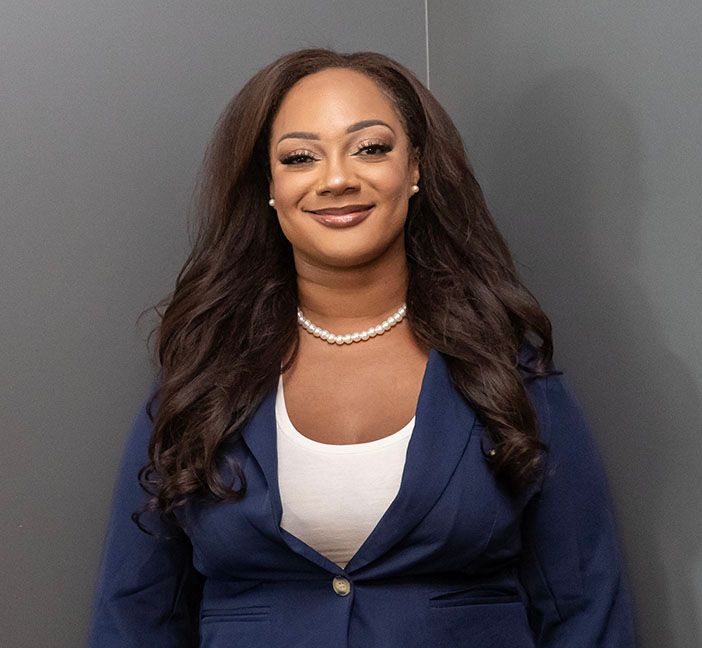 Woman, Ashaki Satomi Scott with long wavy hair smiles, wearing a navy blazer, white top, and pearl necklace. Grey background.