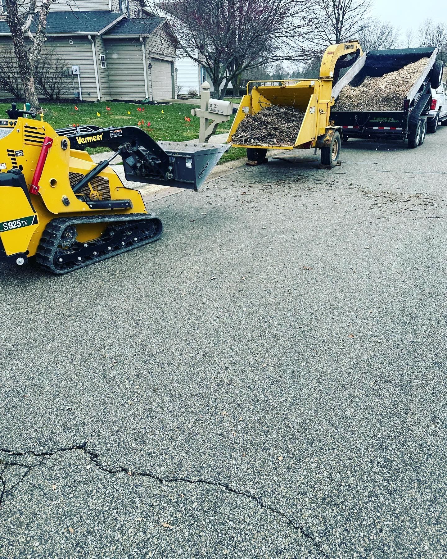 Yellow skid steer loading gravel into a trailer parked on a cracked asphalt road. Houses in the background.