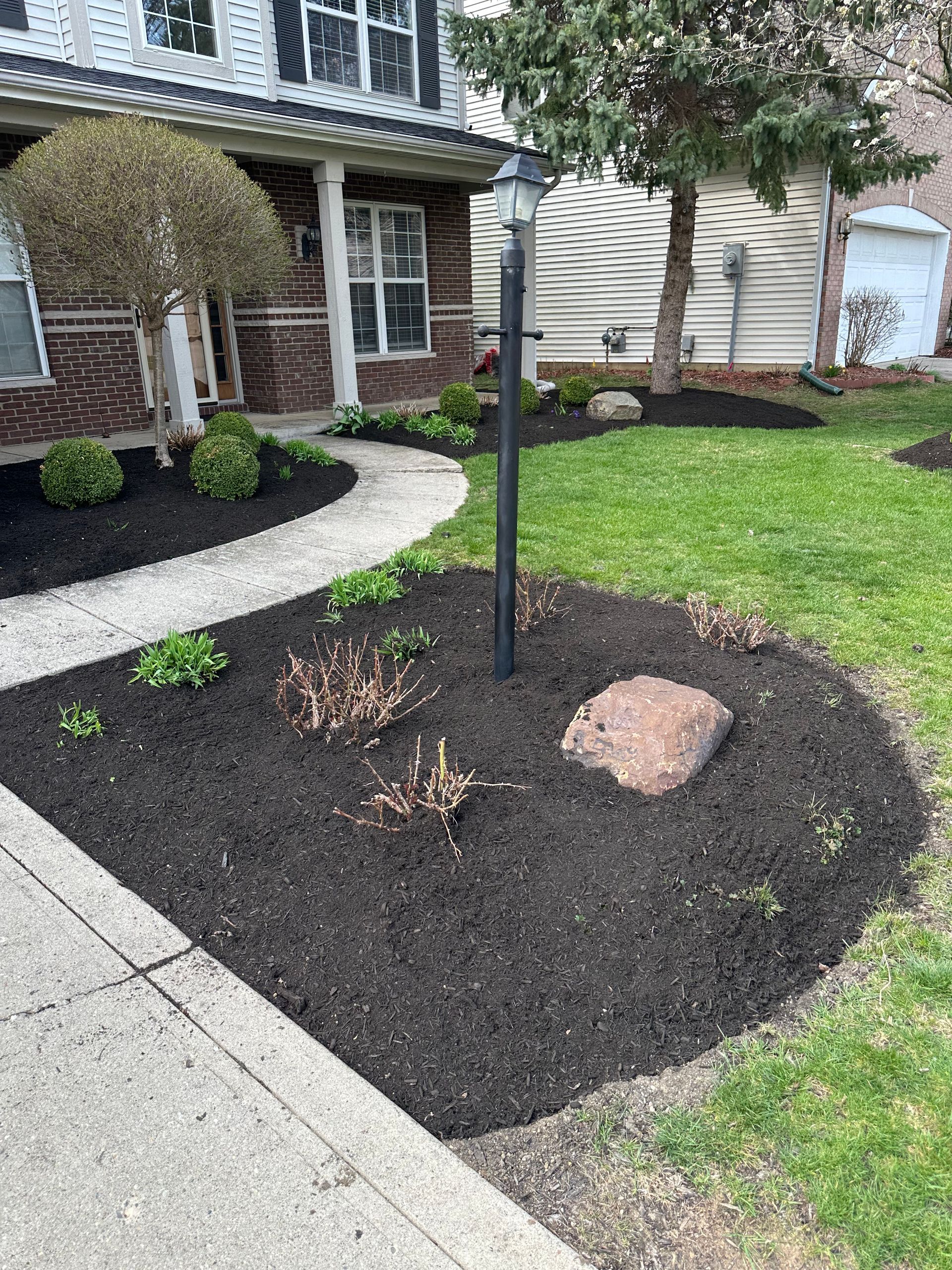 A well-mulched flower bed with a lamppost, rock, and path in front of a house.
