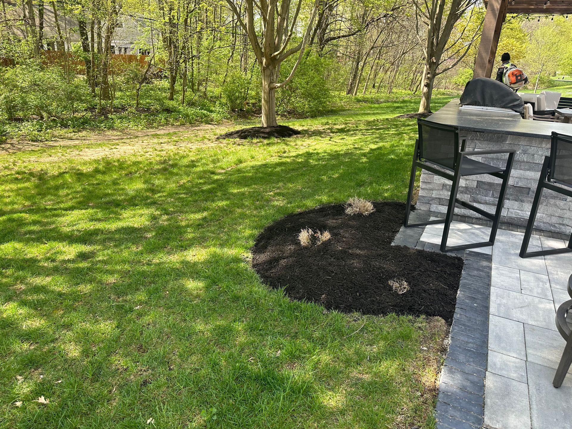 Lush green lawn with a black mulch bed. A stone outdoor kitchen with bar stools.