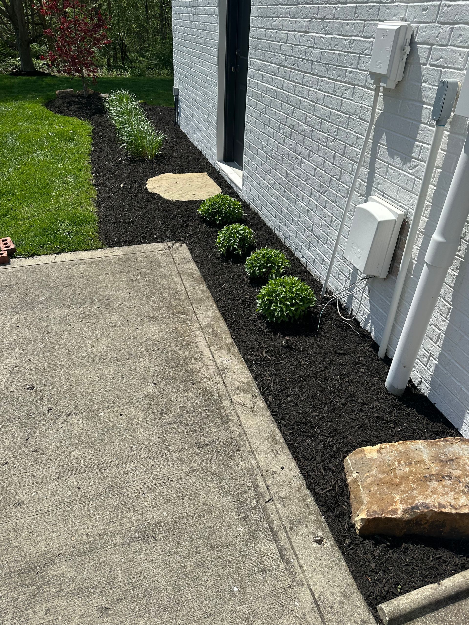 A black mulch flower bed with green plants and a white brick wall.