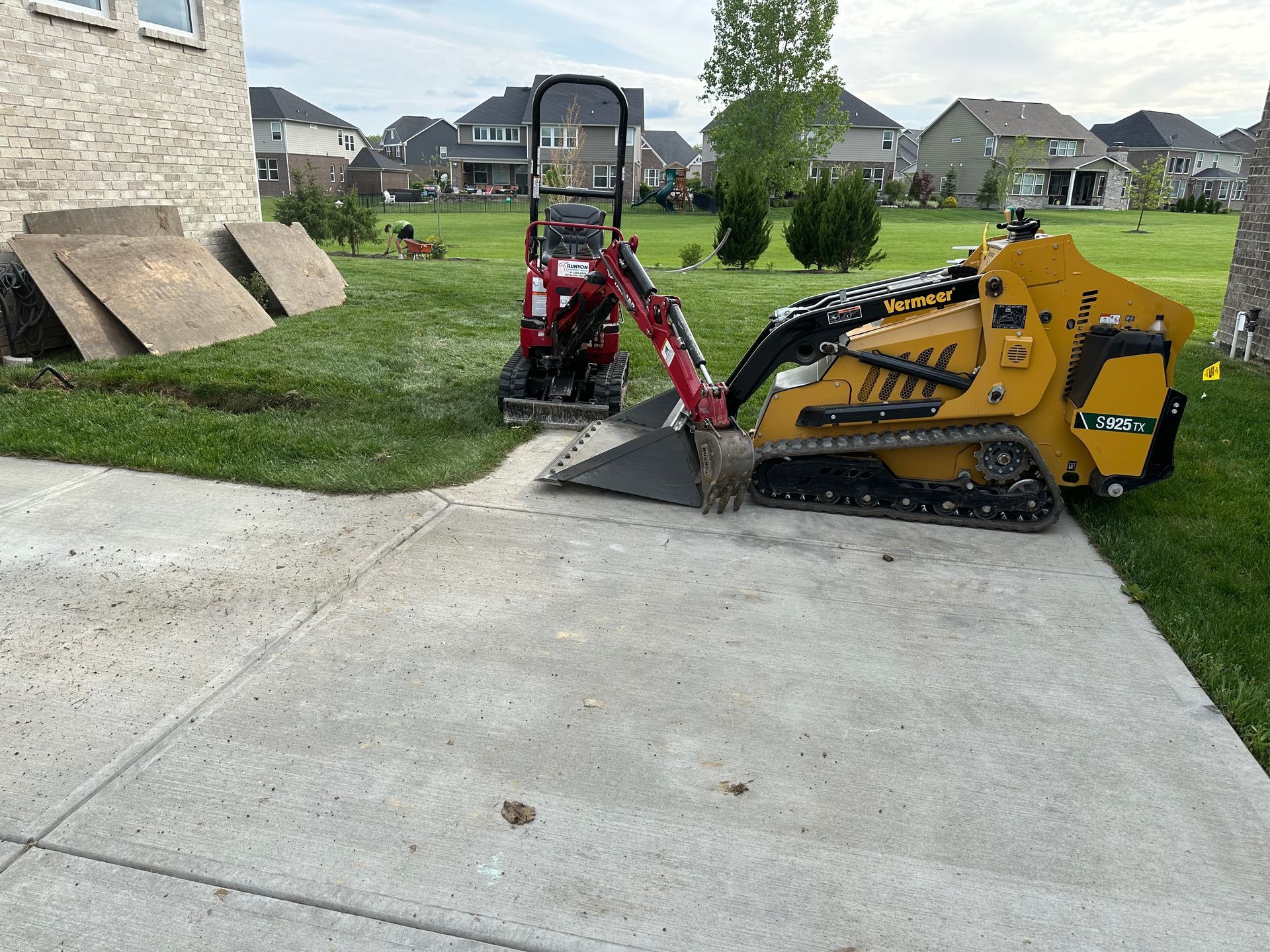 Mini excavator and skid steer on a concrete surface next to grass, near a brick building, working on lawn.