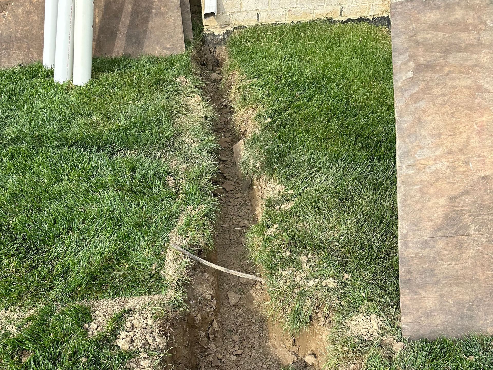 A narrow trench dug in grass, likely for utilities. Brown soil, green grass, near a patio.