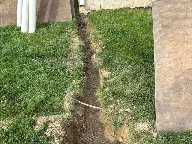 A trench dug in green grass, next to a tan patio and a white PVC pipe.