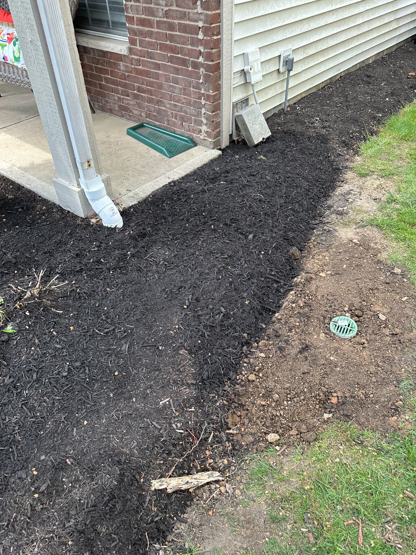 Mulched garden bed next to a house. Dark mulch, green grass, and brick wall.