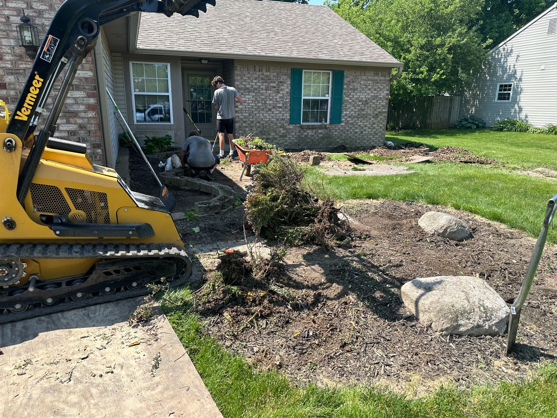 A small, yellow skid steer removing landscape, with two workers in front of a brick house.