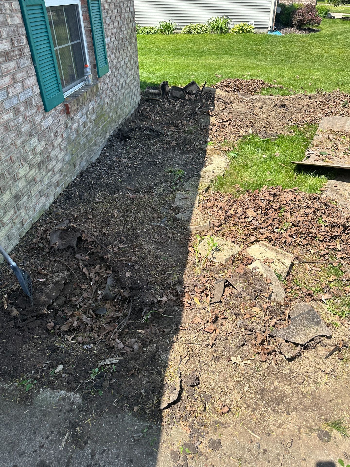 A long, narrow, unkempt flower bed next to a brick house, full of soil and debris.