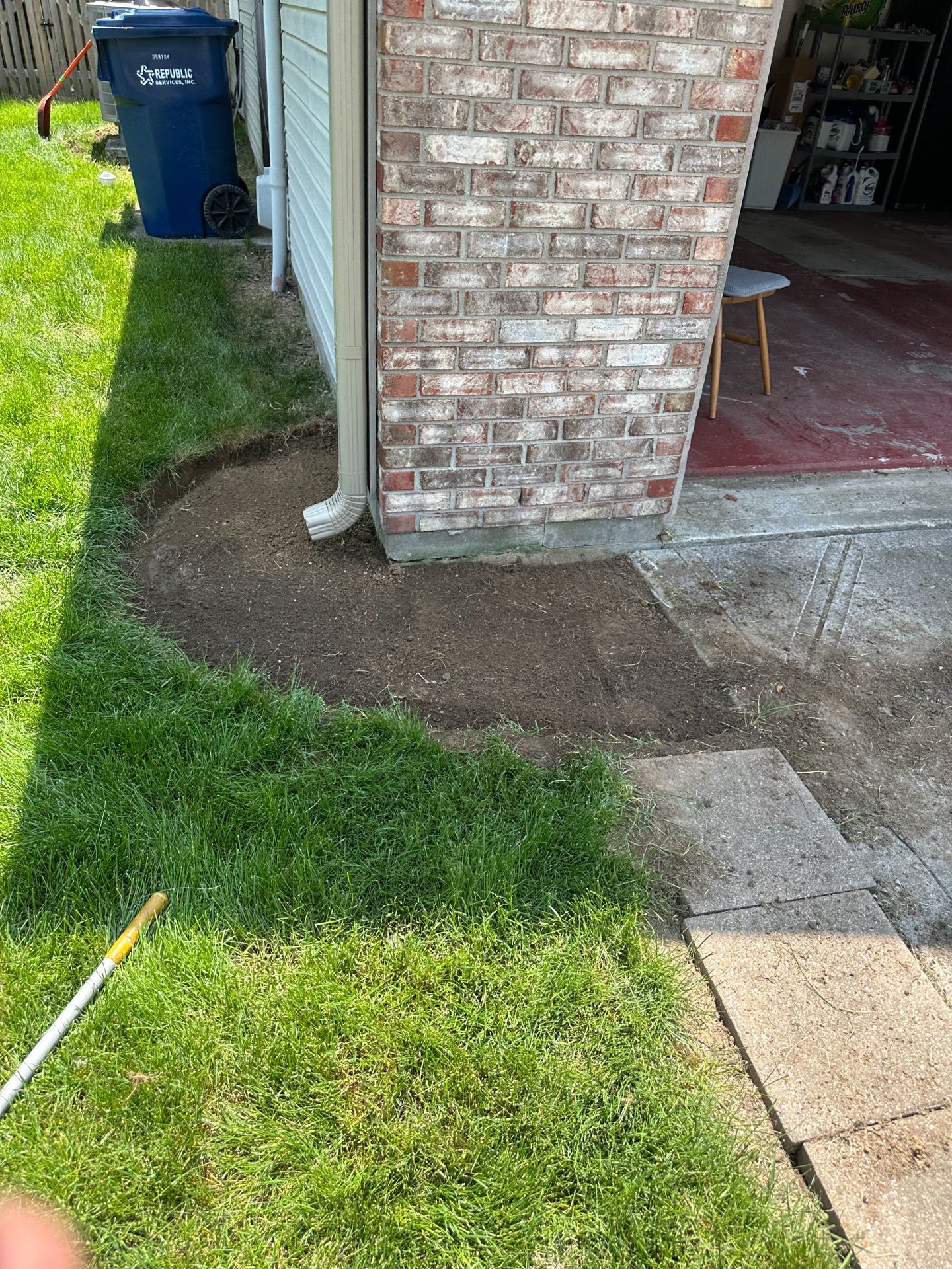 A patch of dirt next to a brick wall, surrounded by green grass. A blue trash bin is in the background.