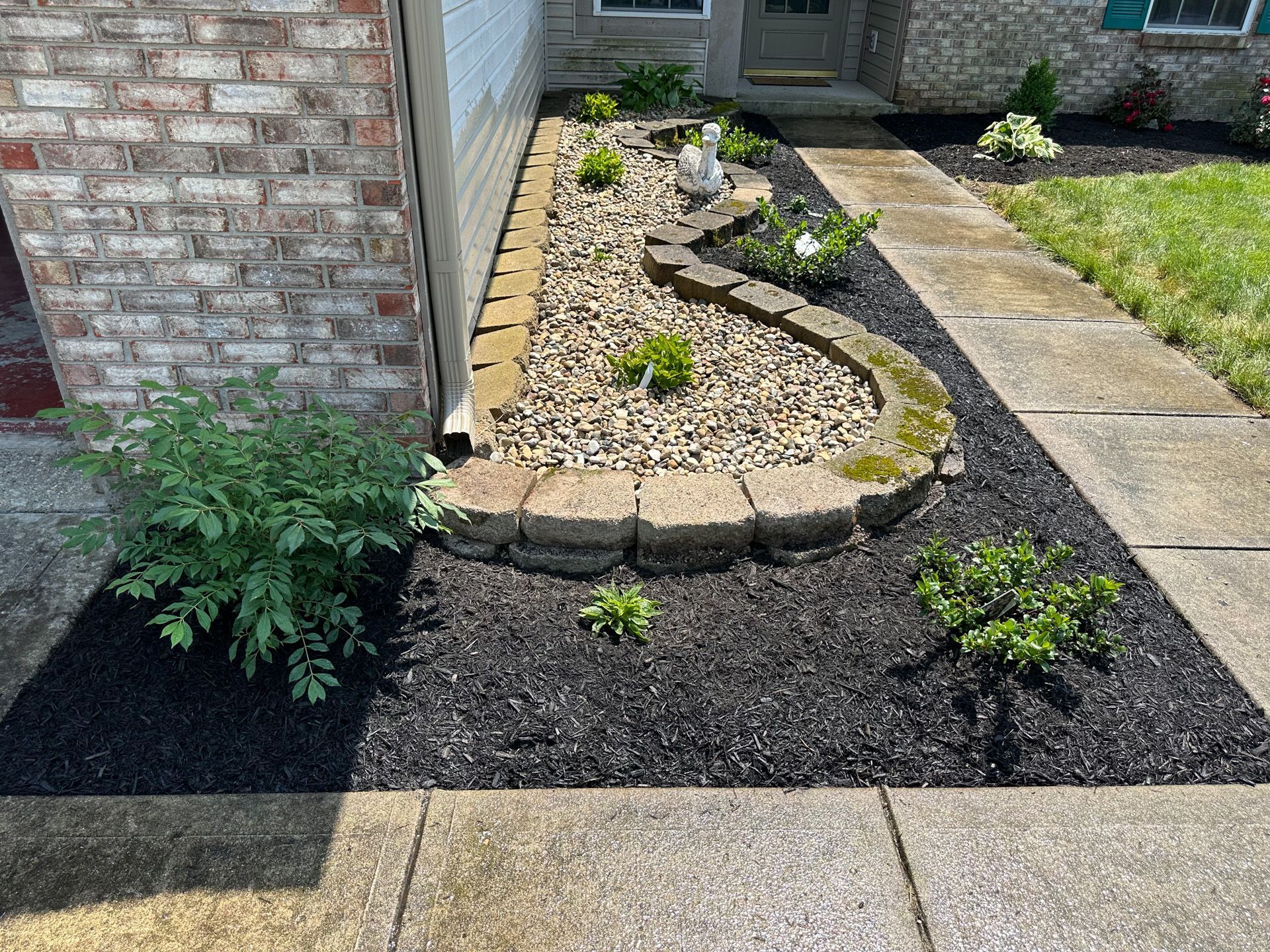 Flower bed with black mulch, stone border, and green plants next to a brick building.