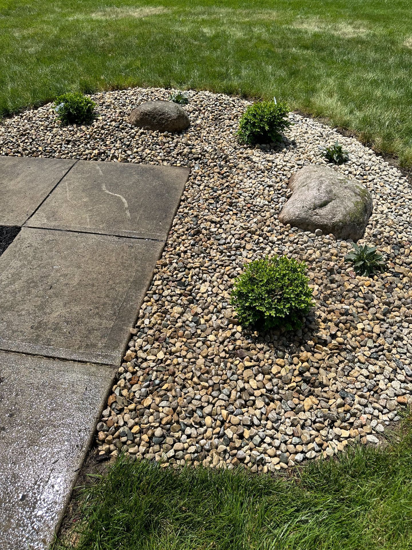Concrete patio next to a rock garden with green bushes and large rocks.