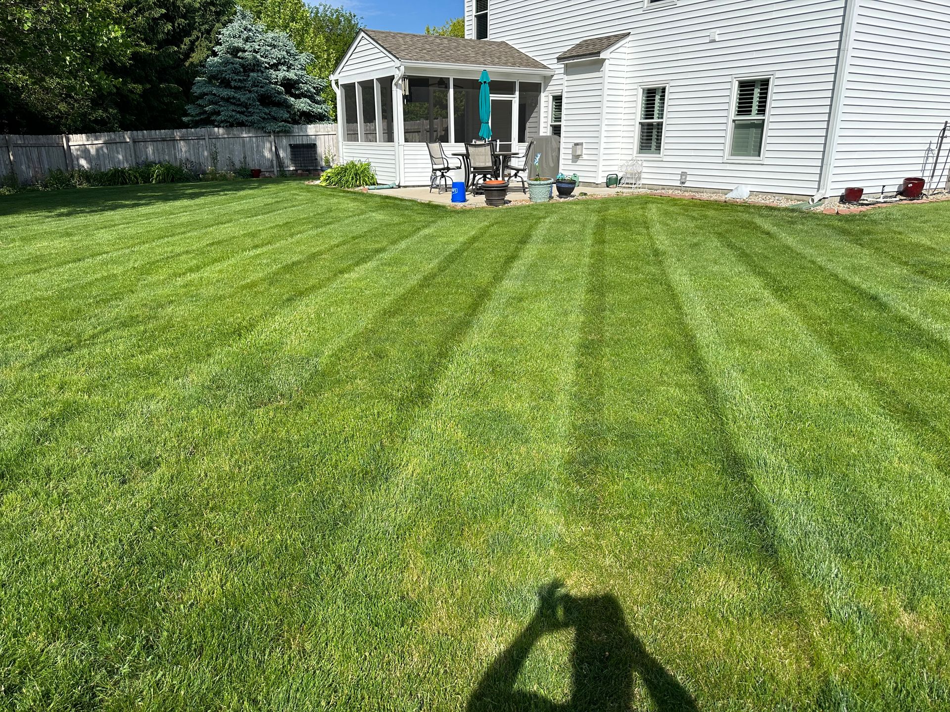 Lawn mowed with stripes, green grass, a white house, screened porch, and shadow of a person taking photo.