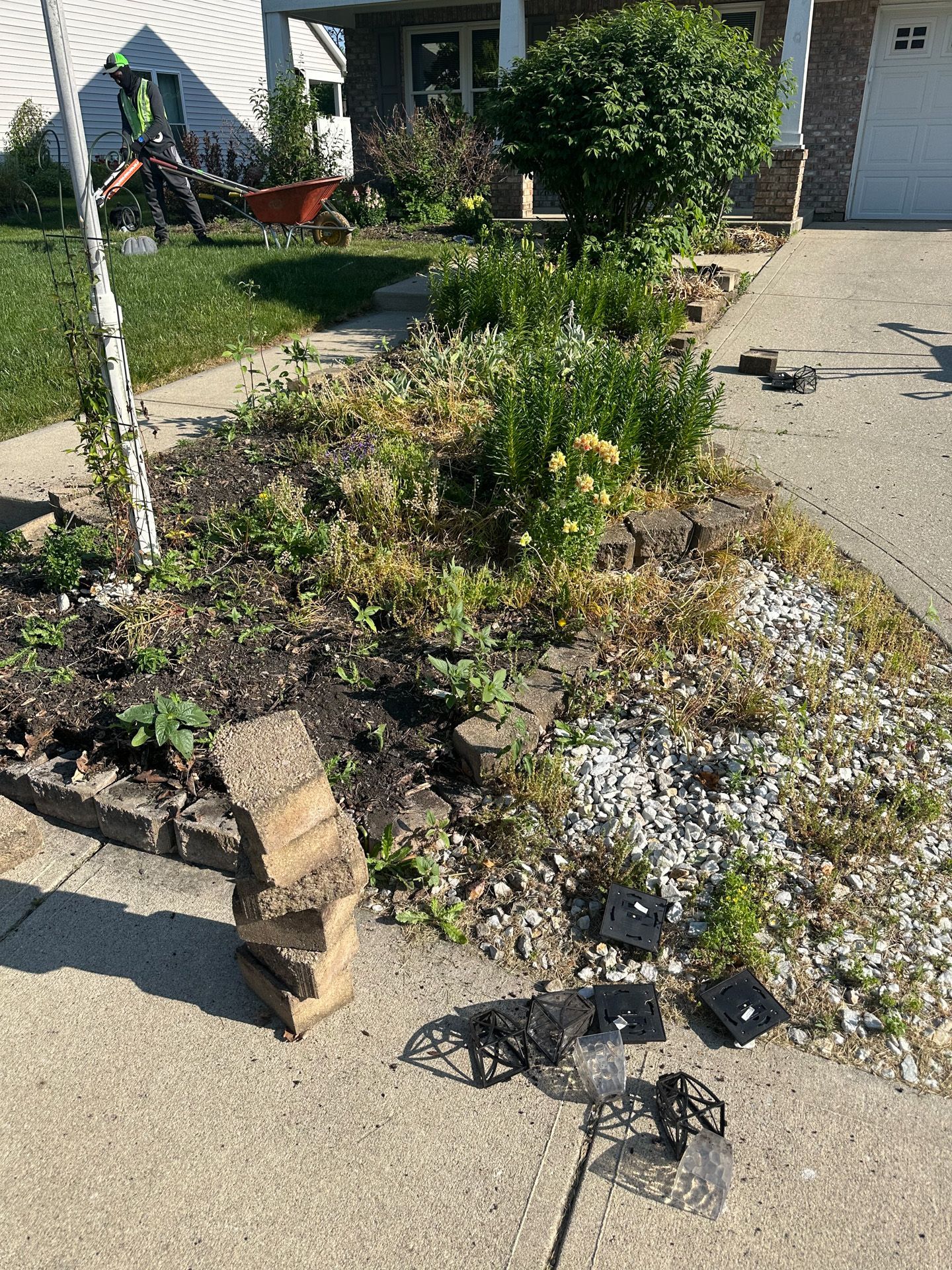 A person is working on a front yard flower bed with stacked bricks and construction materials in the driveway.