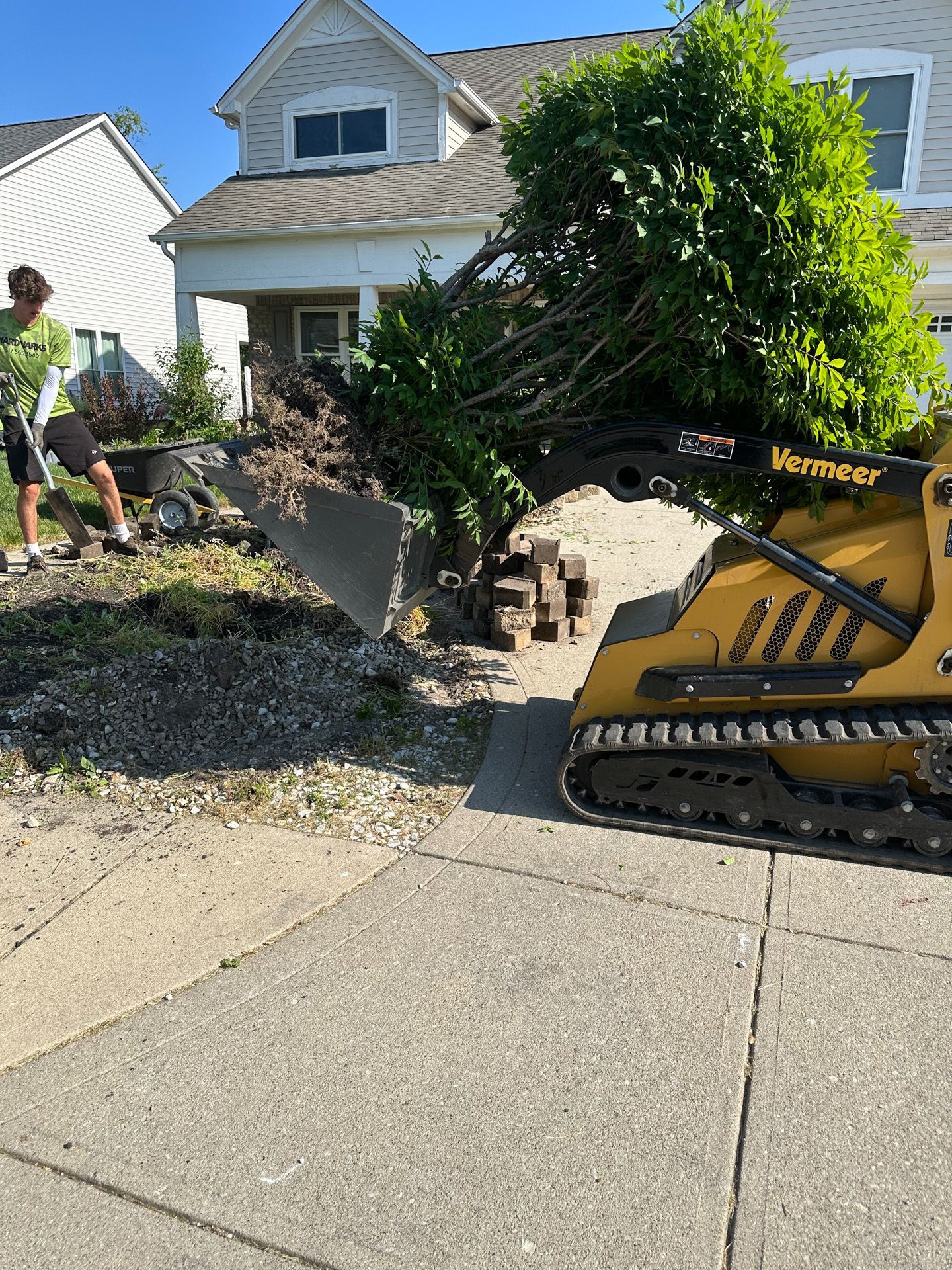 A yellow skid steer removing a large bush near a house; a person helps with a shovel.
