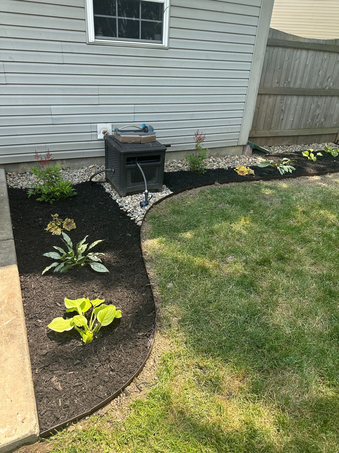 A flowerbed with mulch and plants next to a garage wall and grassy lawn.