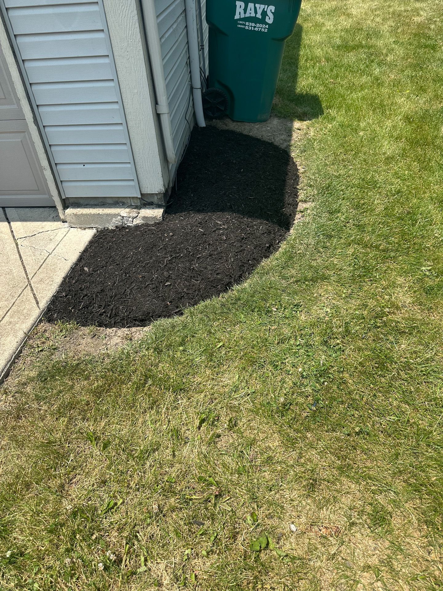 Dark mulch bed next to a white-sided building and green lawn; trash can sits nearby.