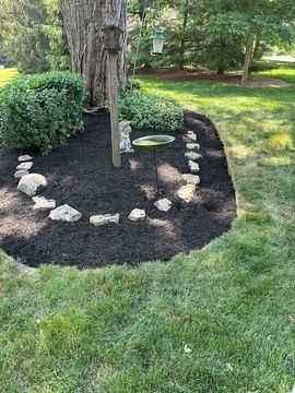 A landscaped garden bed with dark mulch, rocks, birdhouse, and birdbath, surrounding a tree trunk, next to green lawn.