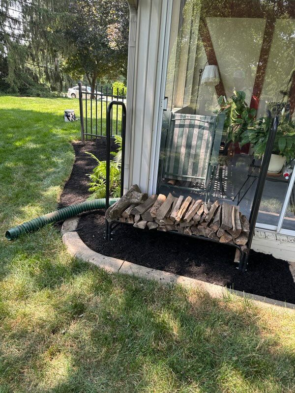 A grassy yard with black mulch border. Firewood on a black rack next to a building column.