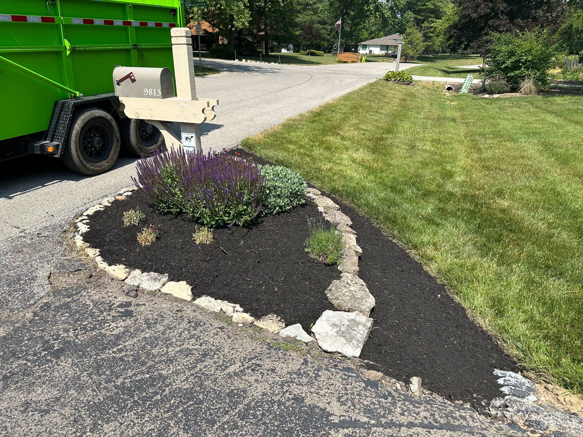Flowerbed with dark mulch, rocks, and plants in a yard next to a driveway, with a green trailer.
