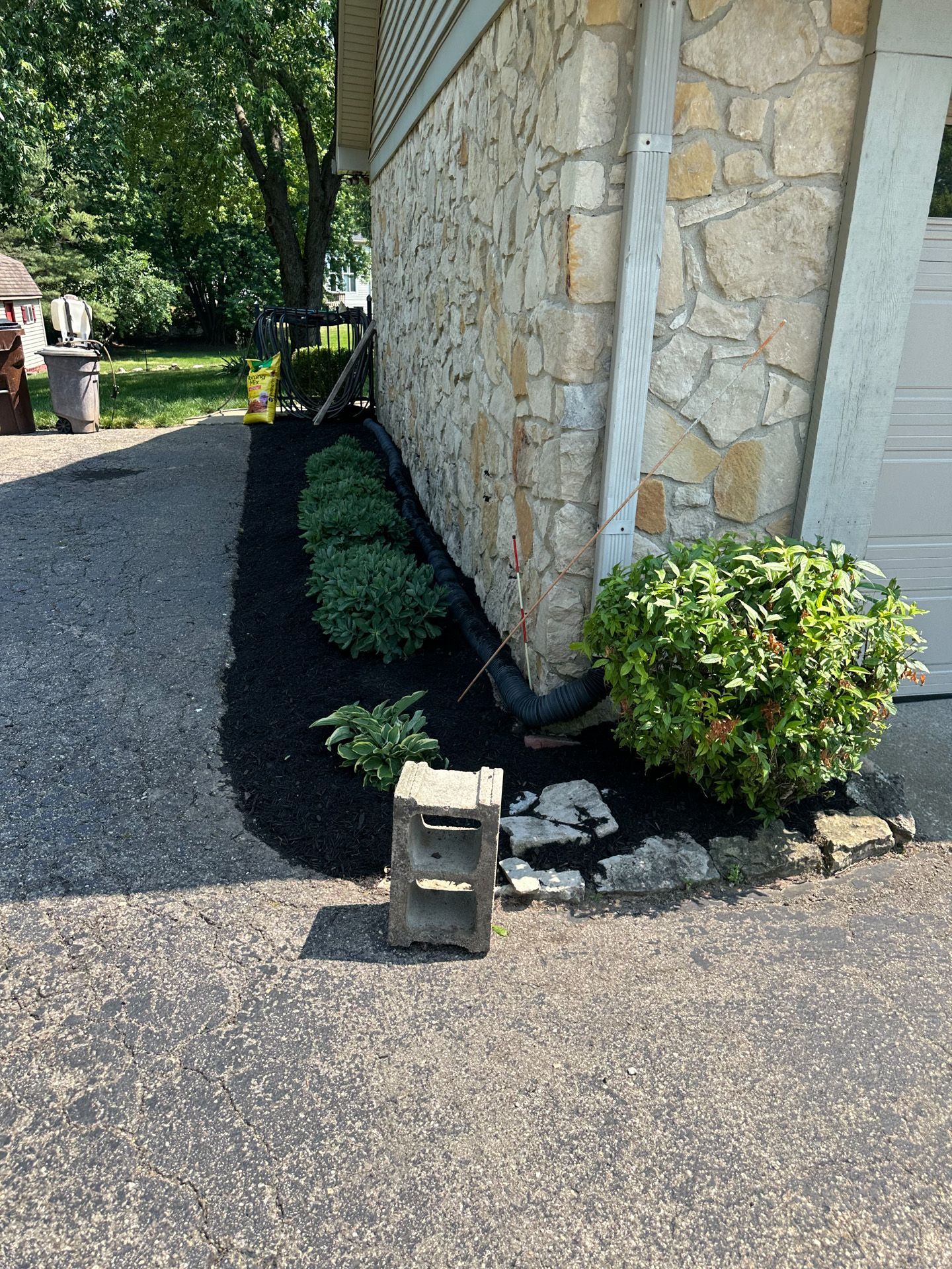 A black-mulched garden bed with green bushes next to a stone-faced building and a paved driveway.