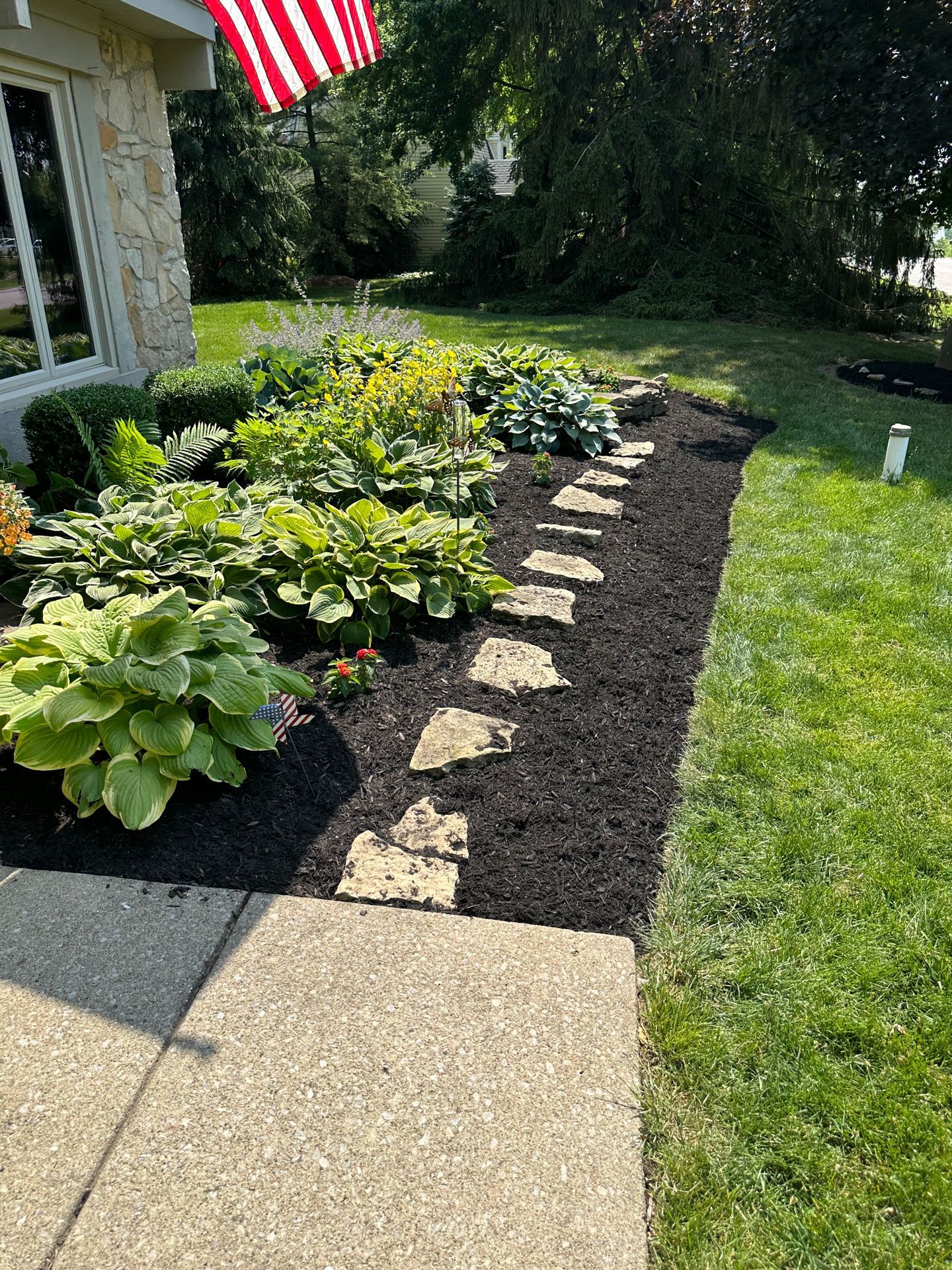 Stone pathway through a flowerbed with lush green plants and black mulch, next to a sidewalk and lawn.