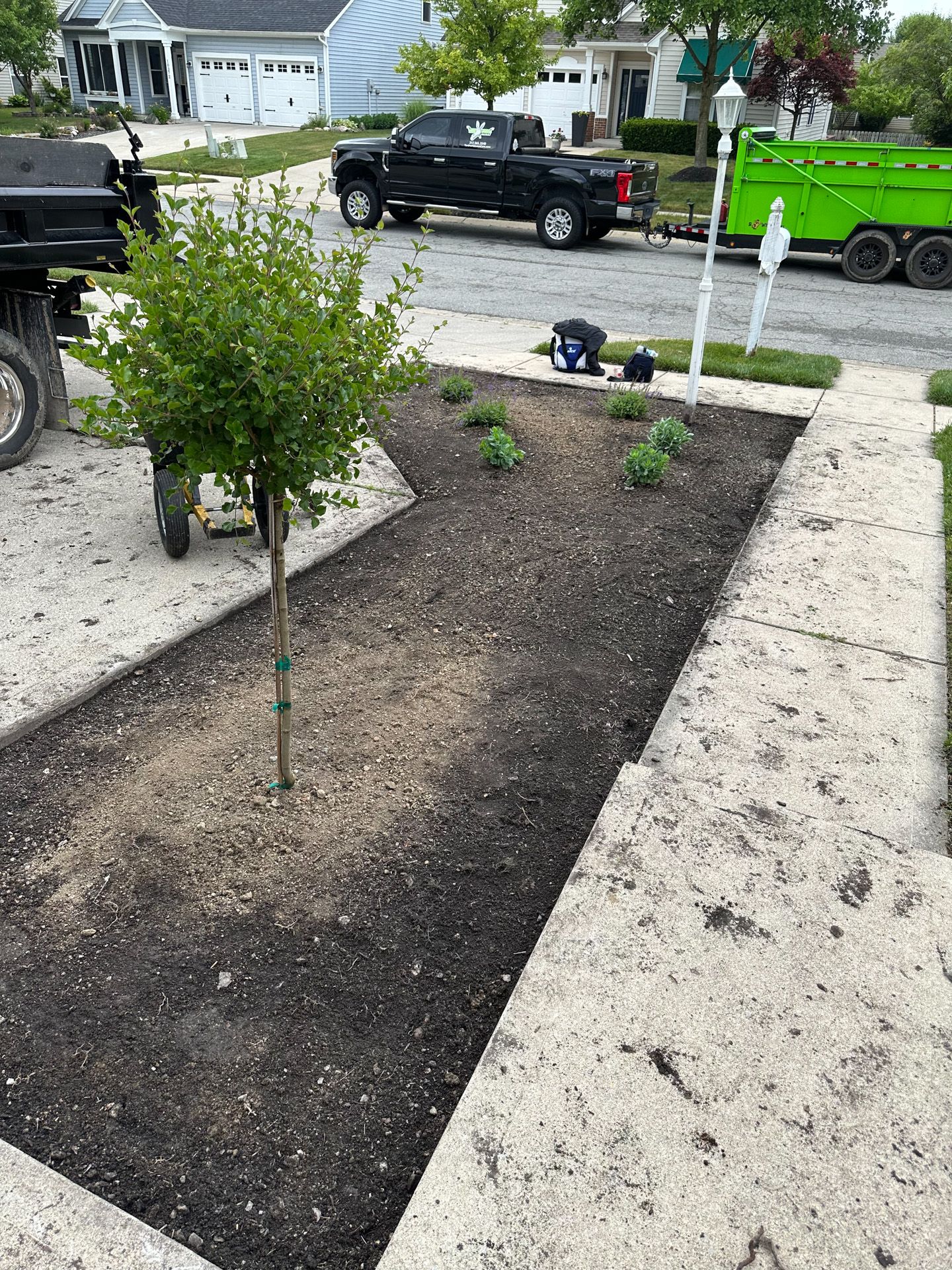 A freshly planted garden bed in front of a house with a small tree and greenery, with a truck in the background.
