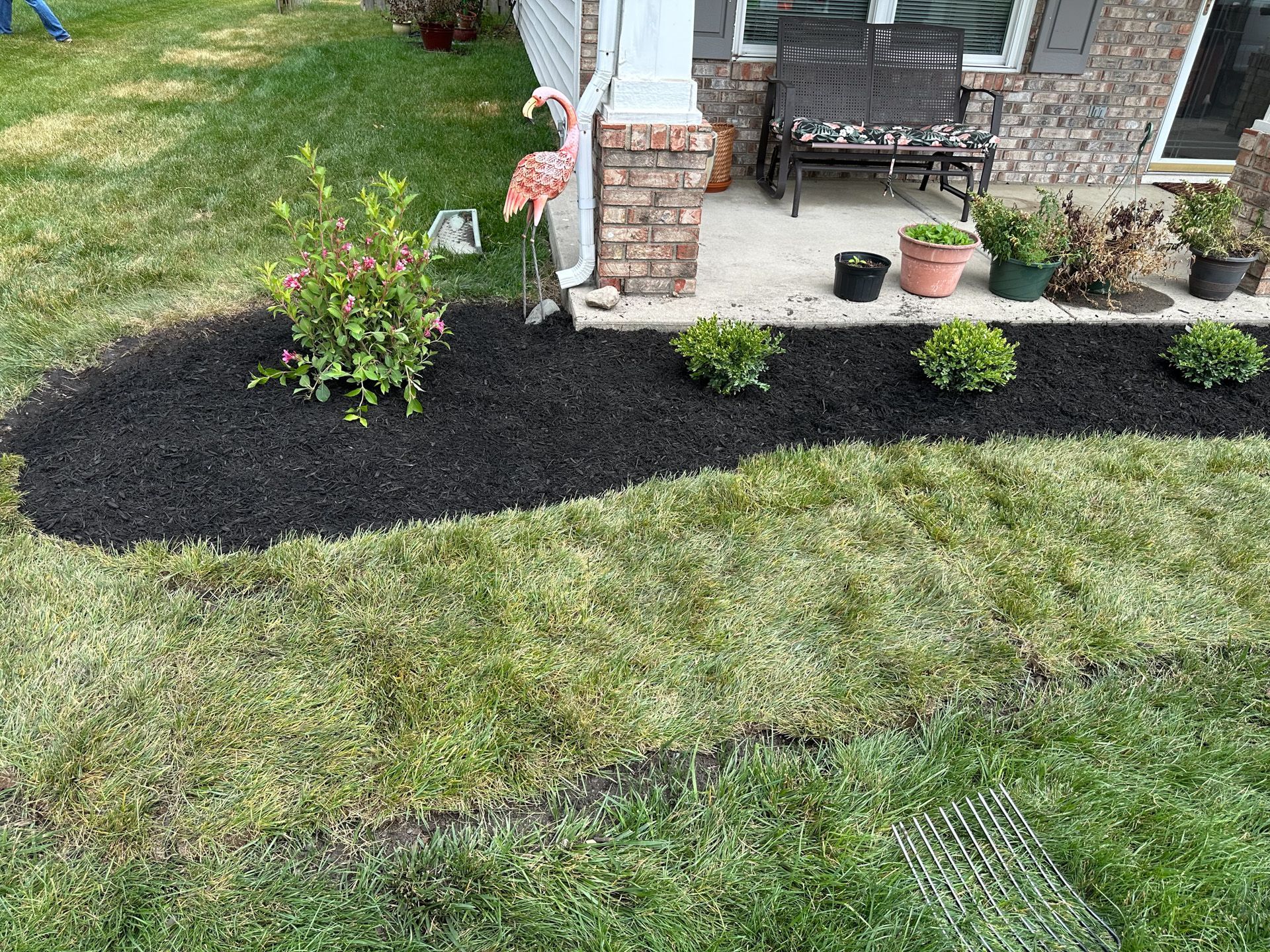 Lush green lawn borders a dark mulch bed with shrubs and a porch with plants.
