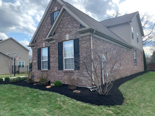 Brick house with black shutters and mulch-lined landscaping against green grass under a cloudy sky.