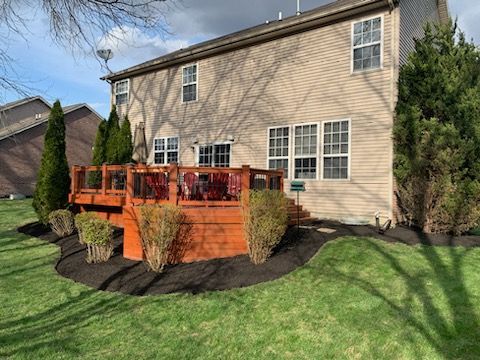 Backyard deck with brown stain, surrounded by mulch beds and lawn. Beige house with white windows.