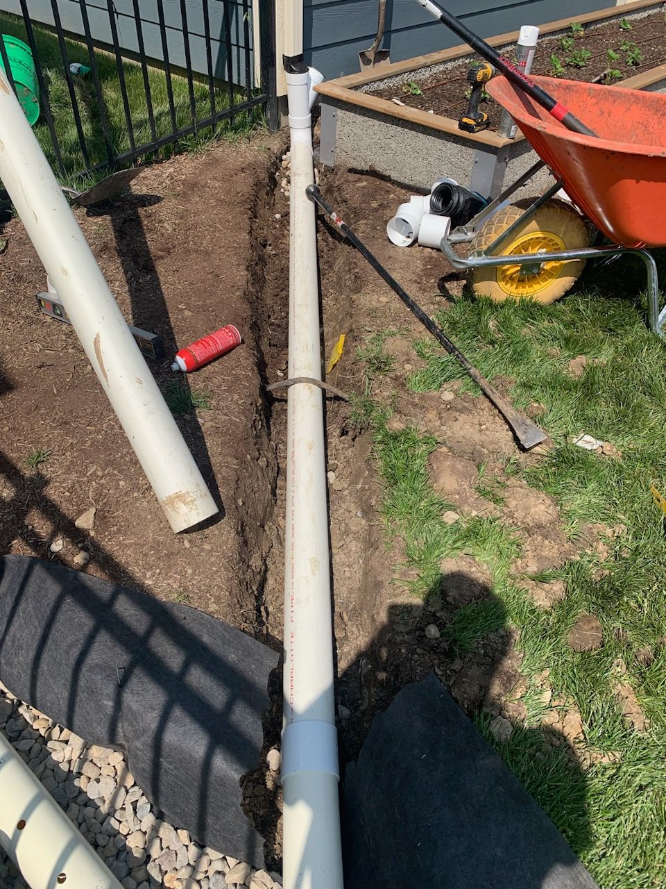 White PVC pipe in a narrow trench, alongside a fence and grassy area, with gardening tools and wheelbarrow nearby.