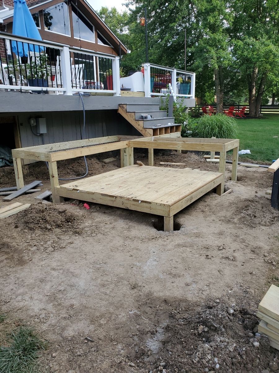 A partially built wooden deck, in a dirt yard, with a larger deck in the background.